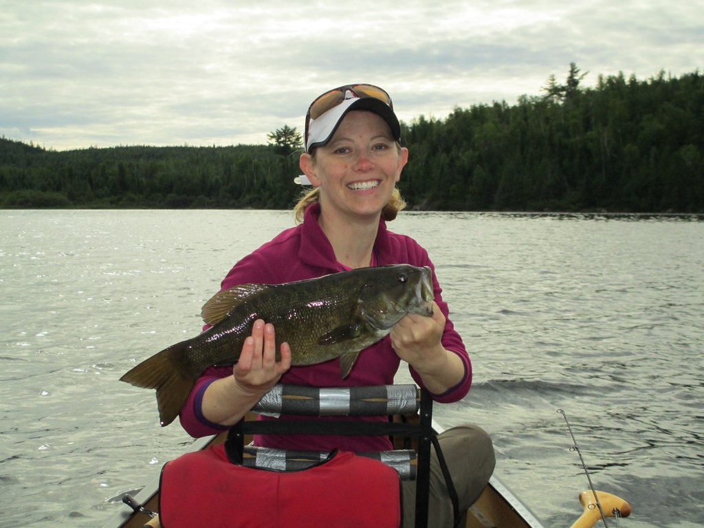 Woman bass angler holding a smallmouth bass from a boat, wearing a pink fishing top